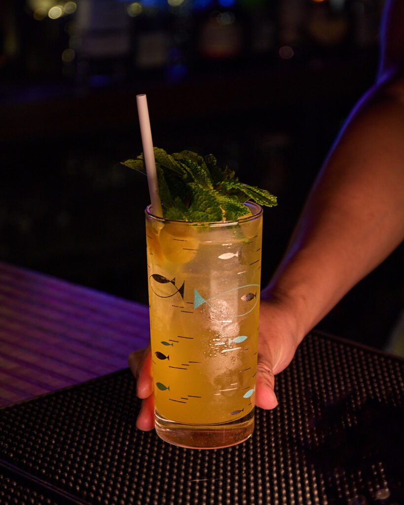 A hand holds a tall cocktail glass with mint and lemon, set on a bar counter.