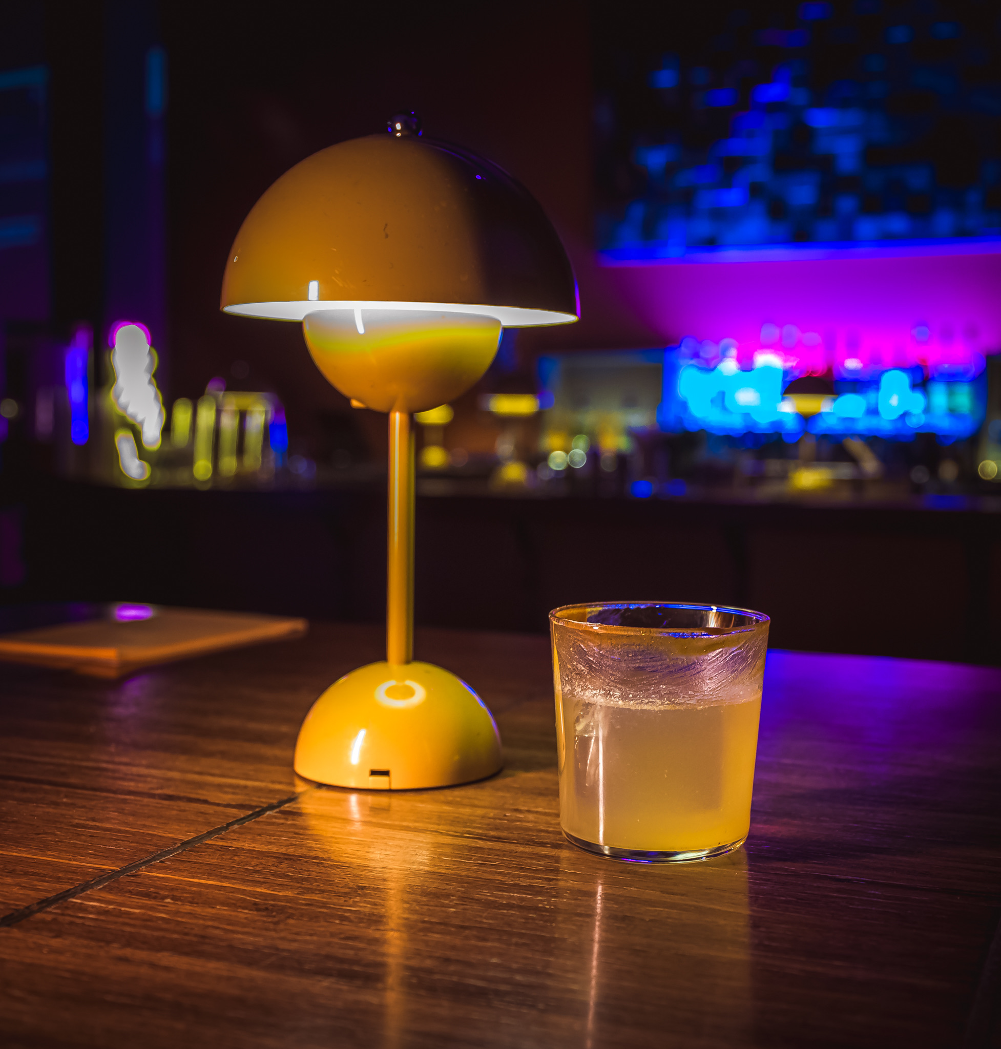 A yellow table lamp and a cocktail glass on a wooden bar, illuminated by colorful lights at the "Play It Cool Pop-Up Bar Event."