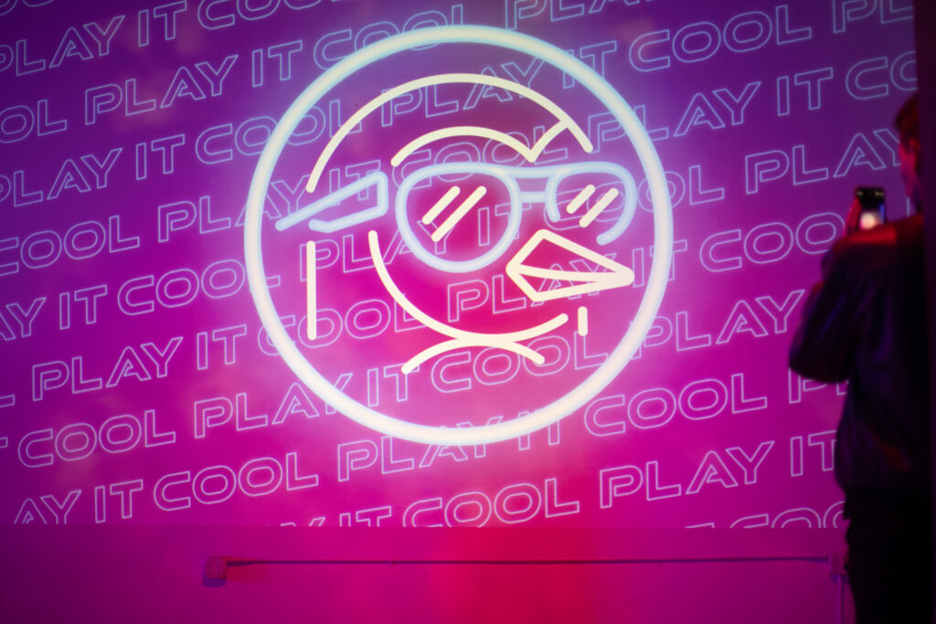 Neon art of a penguin with sunglasses and "PLAY IT COOL" text on a pink background. A person on the right takes a photo.