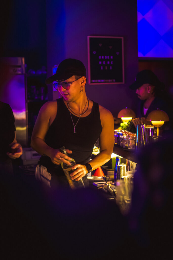 Play It Cool Founder Parker Luthman behind a bar, wearing a black tank top and hat, holding a bottle under dim purple lighting.