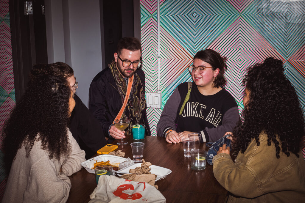 Play It Cool Pop-Up Bar Event: A group of people enjoy drinks and conversation at a table with colorful geometric wall art.