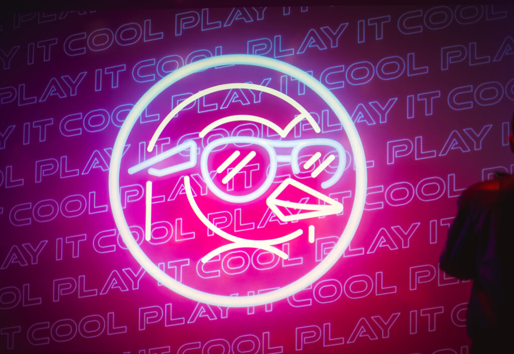Neon "Play It Cool" logo projection featuring a stylized penguin with sunglasses against a vibrant pink background.