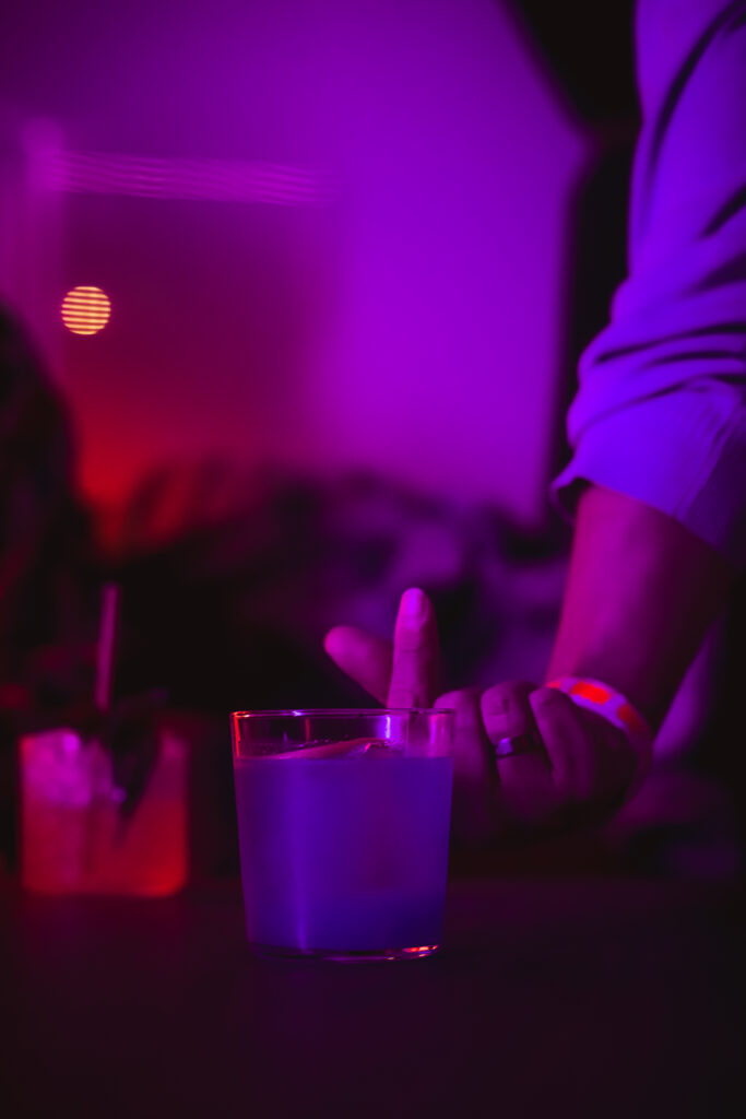 "Play It Cool Pop-Up Bar Event" features a dimly lit scene with vibrant purple and red lighting. A hand with a wristband is pointing near a glowing blue drink on the table.
