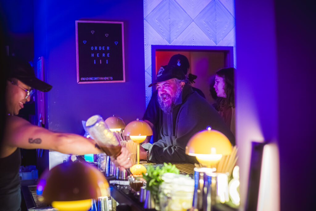 "Play It Cool Pop-Up Bar Event" with a smiling bartender pouring a drink, and a bearded man in a cap enjoying the atmosphere.