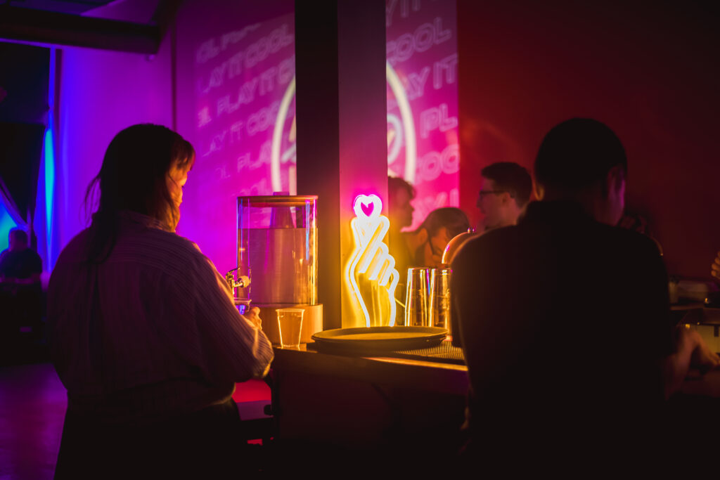 Play It Cool Pop-Up Bar event featuring a glowing finger heart neon sign, with patrons enjoying the vibrant atmosphere.