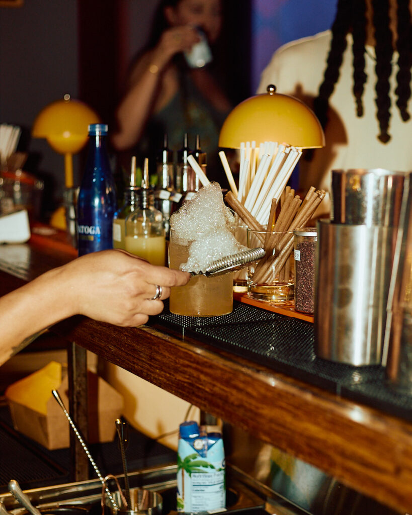A hand garnishes a cocktail with foam at a bar, surrounded by bottles and straws.
