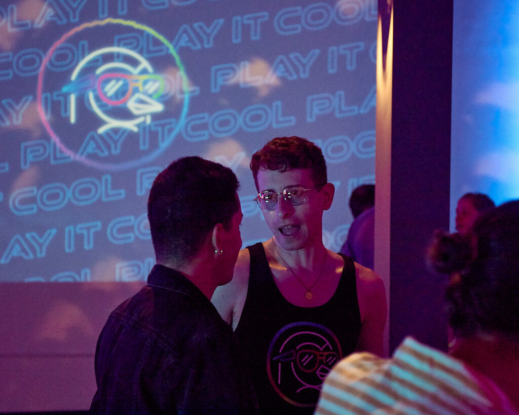 Two people converse in a vibrant setting with a colorful "Play It Cool" backdrop.