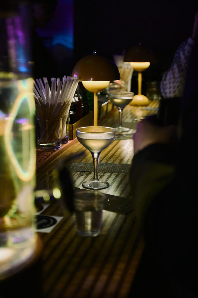 A dimly lit bar with cocktails, illuminated lamps, and a person holding a drink.
