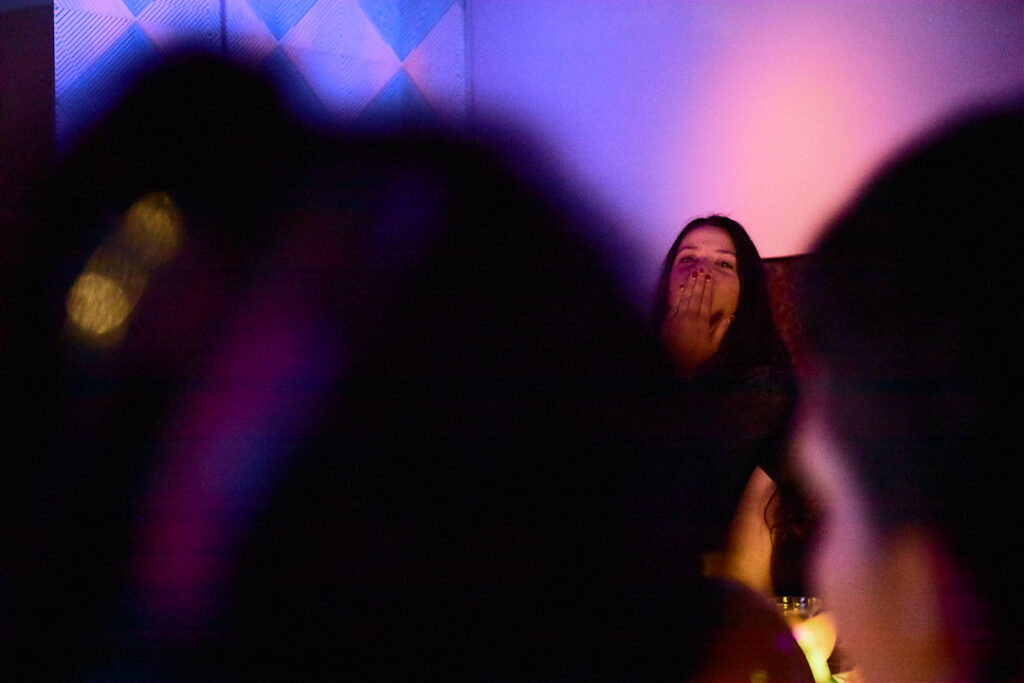 A woman sits in a dimly lit space, blowing a kiss while surrounded by blurred figures.