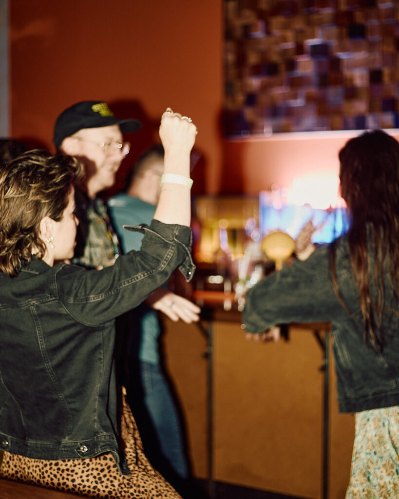 People dance and celebrate at a lively bar, with drinks on the counter and colorful lights in the background.