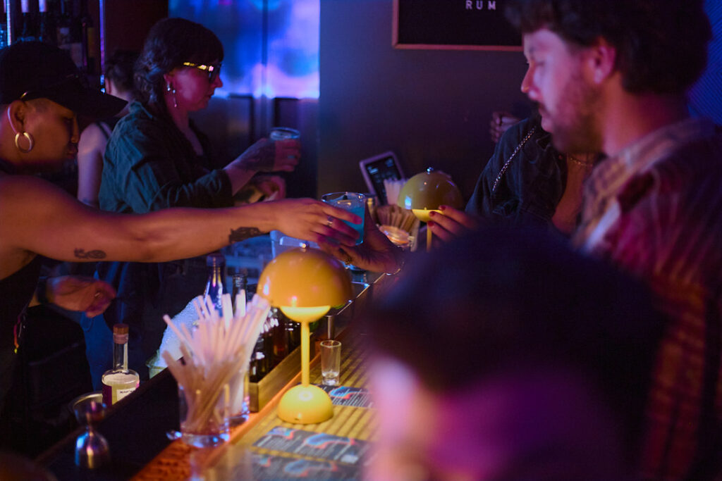 Two people exchange drinks at a bar, colorful lighting and decor create a lively atmosphere.