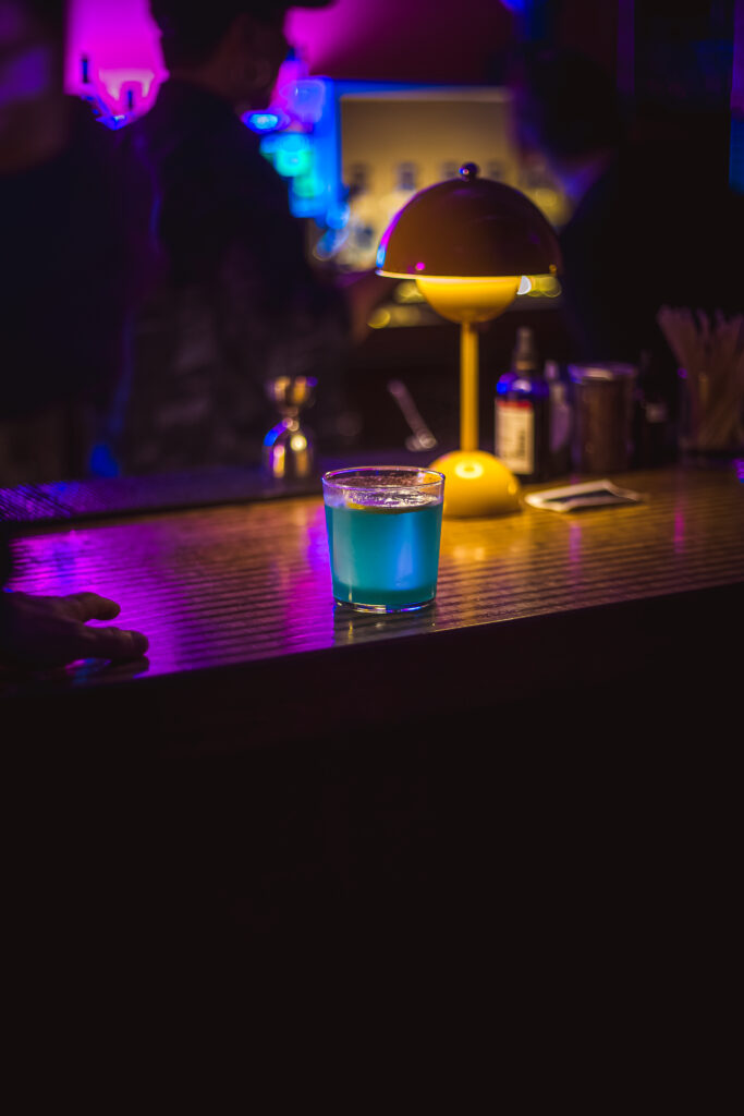 Blue cocktail on a dimly lit bar counter at "Craft Cocktail Play It Cool Pop-Up Bar Event," with a yellow lamp nearby.